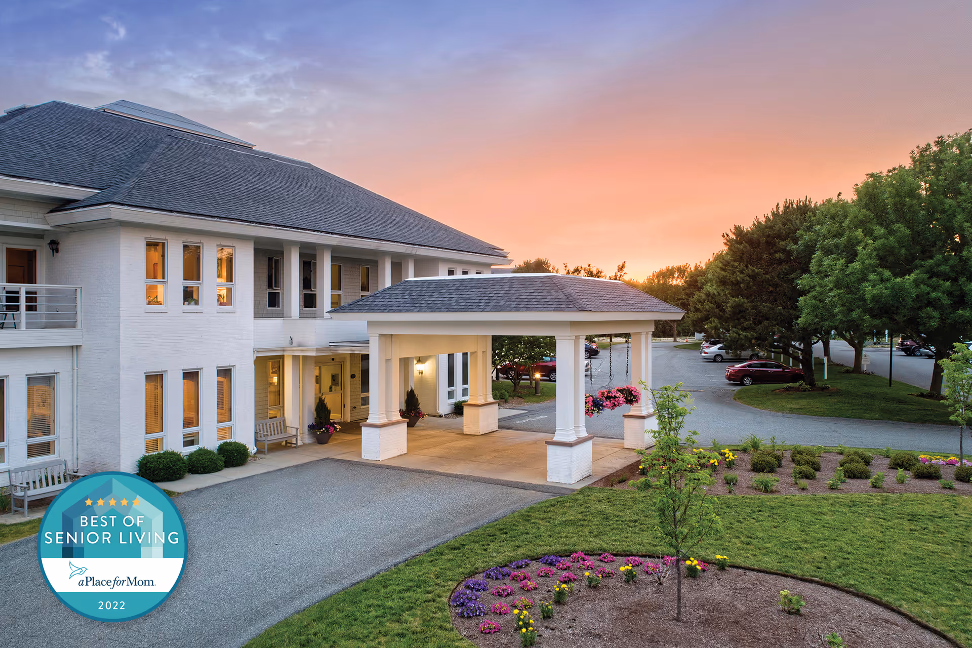 Front entrance of a senior living building with a covered porte-cochere, landscaped flowerbeds and a colorful sunset sky.