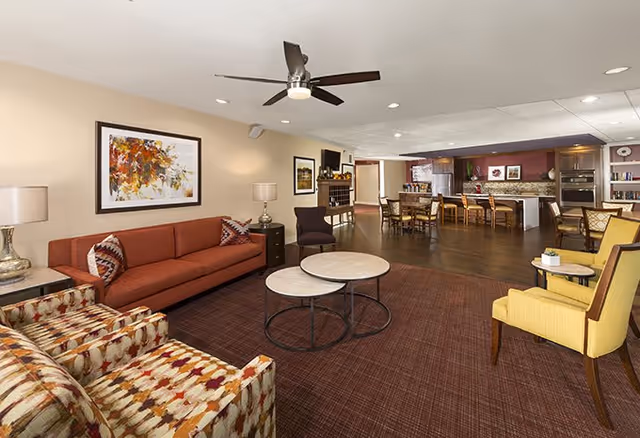 A spacious senior living common area featuring a red sofa, patterned armchairs, two round nested coffee tables, yellow armchairs, and a ceiling fan. The room has warm lighting, artwork on the walls, and an open view into a dining area and kitchen with wooden floors and cabinetry.
