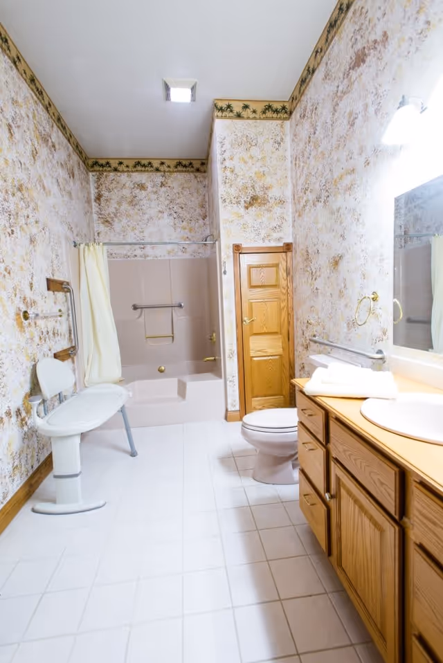 A senior-friendly bathroom featuring a walk-in bathtub with grab bars and a shower chair. The room has floral-patterned wallpaper with a palm tree border near the ceiling, a wooden vanity with a sink and folded towels, a toilet, and a large mirror above the sink. The floor is tiled in white.