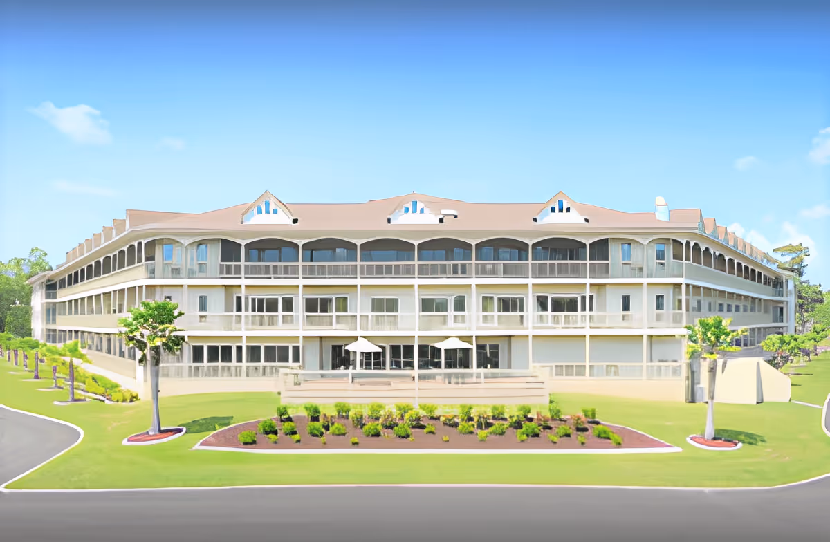 Front exterior of a three-story senior living building with wraparound balconies, manicured lawn and landscaped beds under a clear sky.