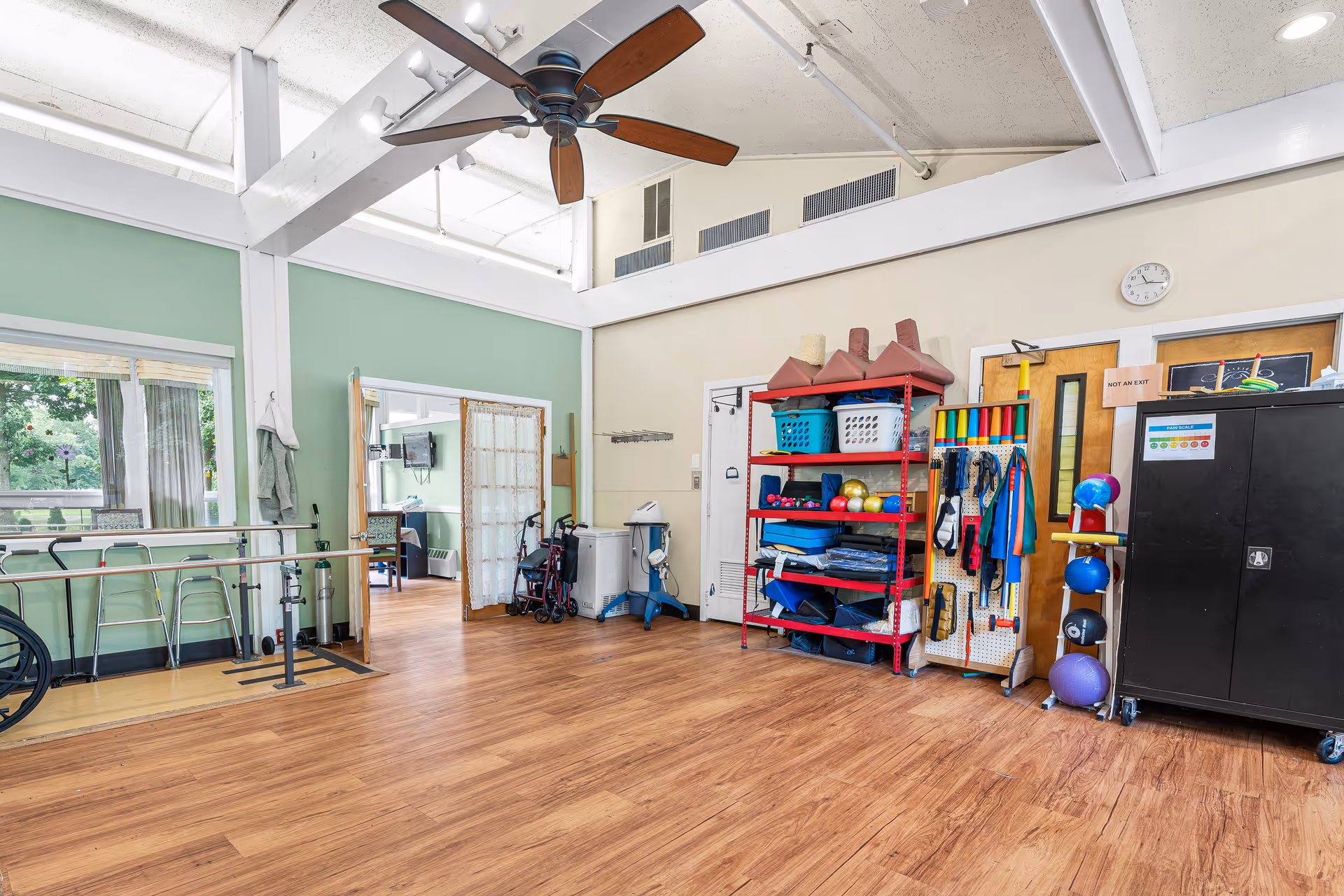 A spacious rehabilitation or therapy room with wooden flooring, a ceiling fan, and large windows letting in natural light. The room contains parallel bars for walking exercises, a red shelving unit with various therapy equipment like exercise balls, weights, and resistance bands, and a black cabinet. There is a doorway leading to another room with a wheelchair visible inside.