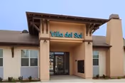 Front exterior view of a single-story building with a covered entrance. The building has beige and light gray walls, a brown roof, and a sign above the entrance that reads 'Villa del Sol'.