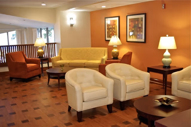 A cozy living room area with a yellow sofa, two orange armchairs, and two white tufted armchairs arranged around dark wooden coffee tables. The room has warm orange walls with two framed abstract artworks and three table lamps providing soft lighting. Large windows with white curtains allow natural light to enter the space.