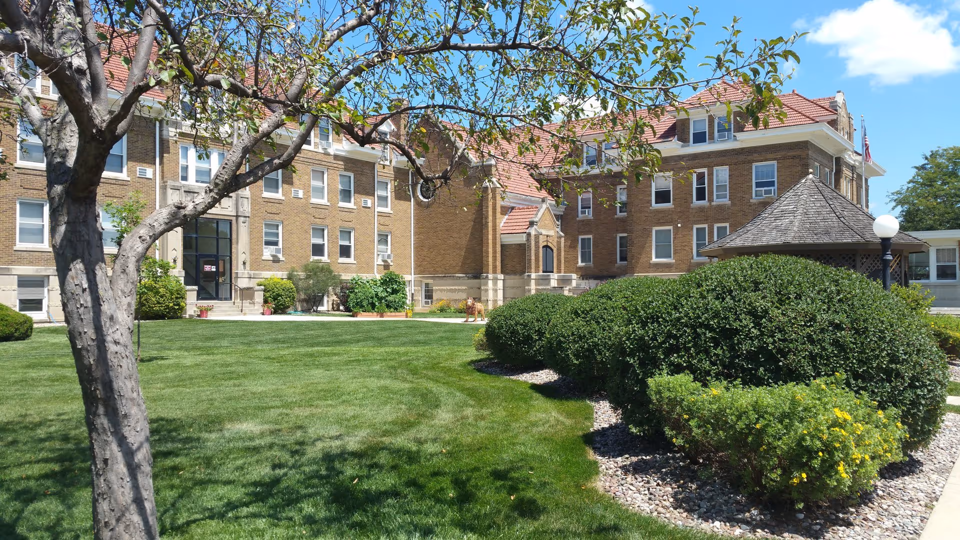Front exterior of a multi-story brick building with a manicured lawn, bushes, a tree, and a small gazebo.
