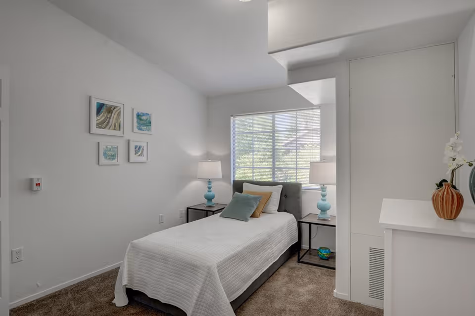 Small bright bedroom with a single bed, two bedside tables and lamps, a window, and framed wall art.