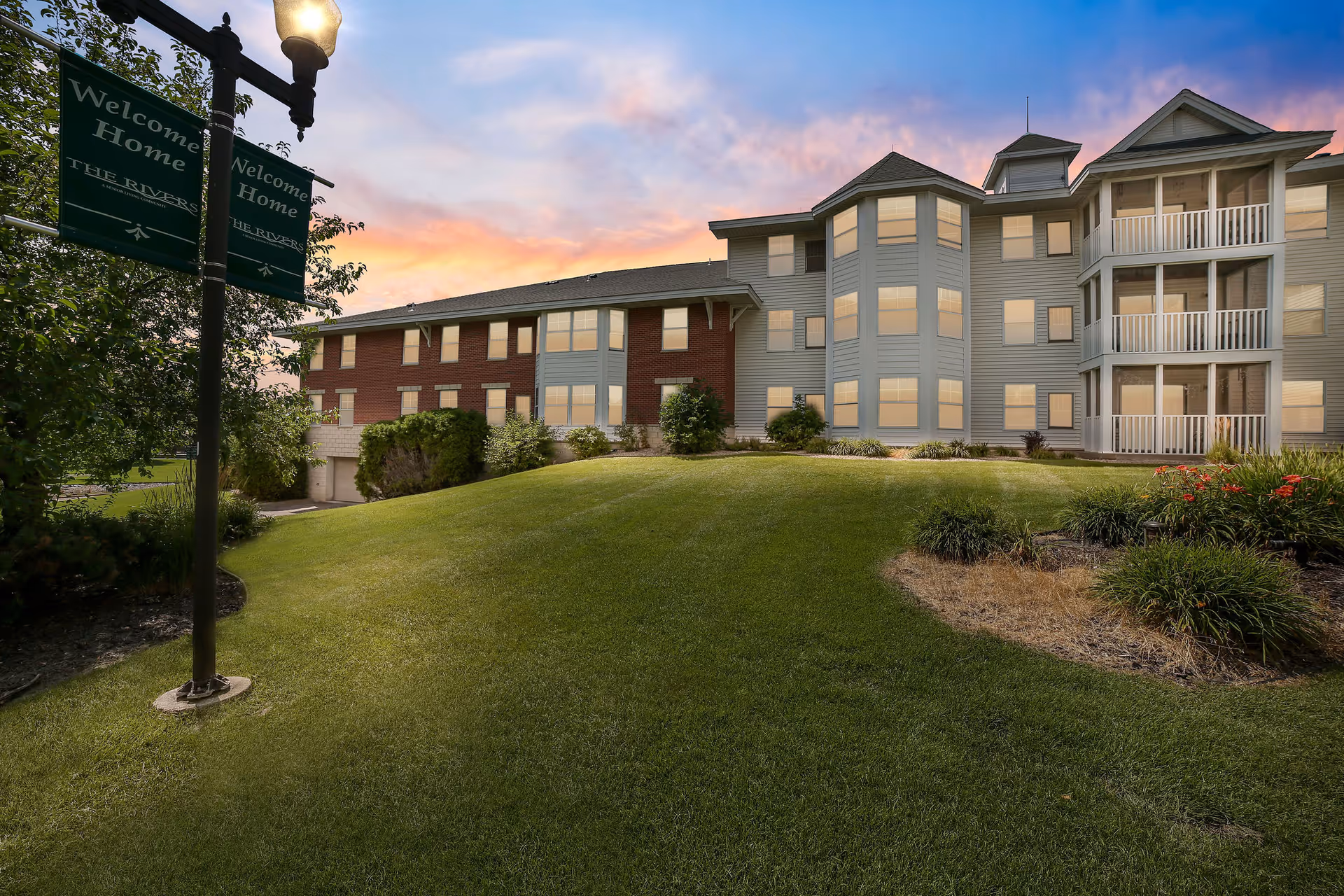 Exterior view of The Rivers Retirement Community building at sunset with a well-maintained lawn and landscaping. A lamppost with green banners reading 'Welcome Home THE RIVERS' is visible on the left side.