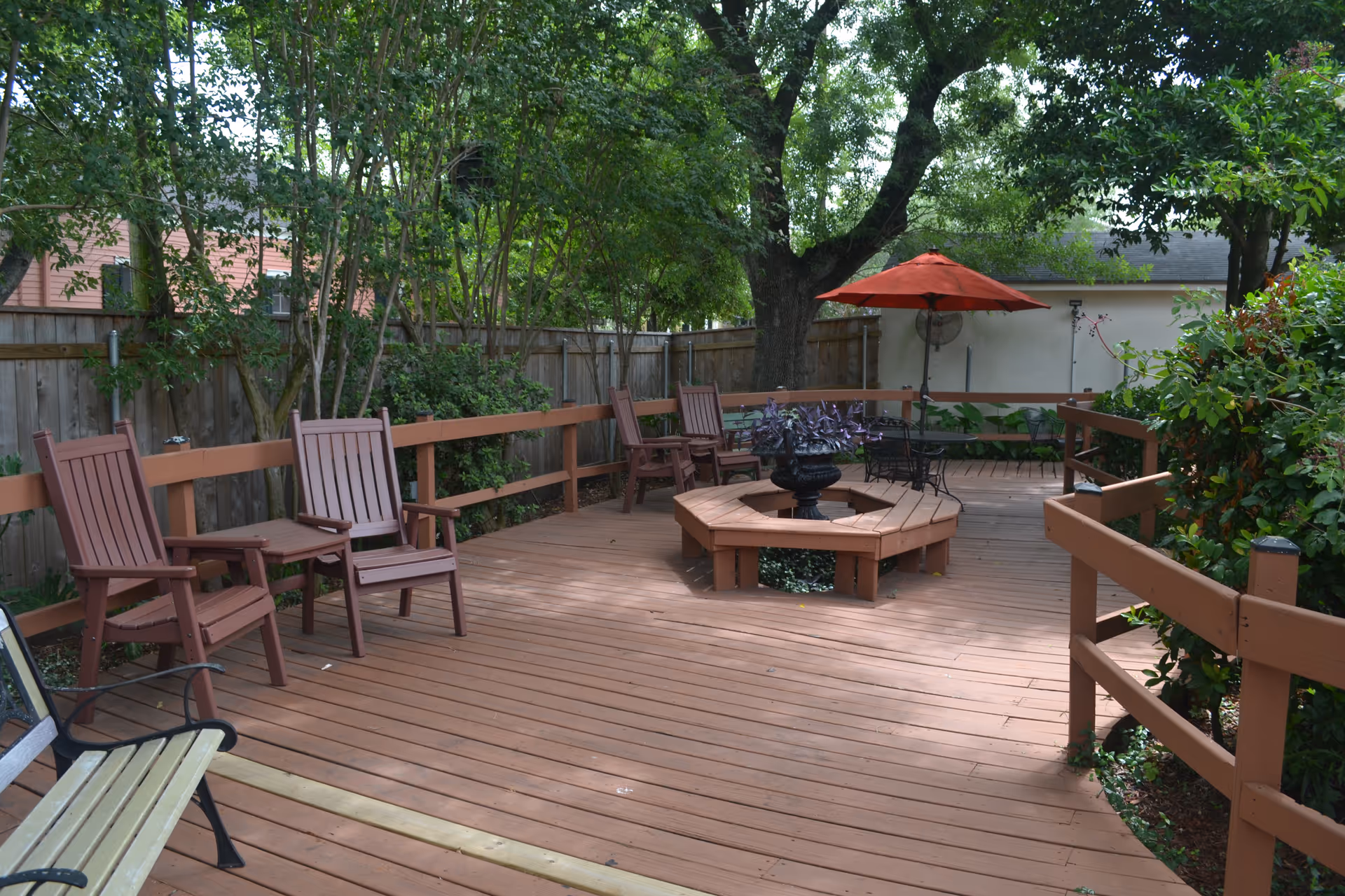 A wooden outdoor deck area with several brown chairs arranged along the railing, a central hexagonal bench surrounding a planter with purple plants, and a red patio umbrella providing shade. The deck is surrounded by trees and greenery, creating a peaceful garden setting.