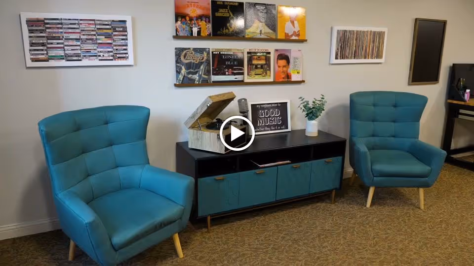 A cozy seating area with two teal upholstered armchairs facing a black and teal cabinet. On top of the cabinet is a vintage-style record player, a framed sign that reads 'my neighbors listen to GOOD MUSIC whether they like it or not,' a microphone, and a small potted plant. The wall behind features shelves displaying vinyl record covers and framed artwork of cassette tapes and vinyl records.