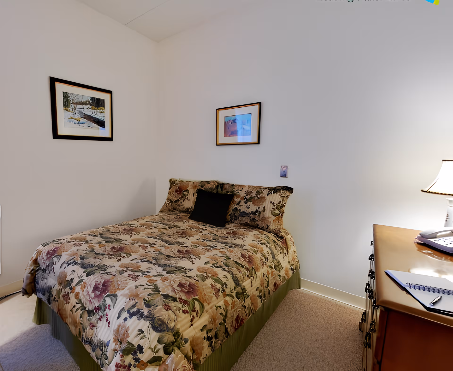 A bedroom with a floral-patterned bedspread on a bed, a black pillow, two framed pictures on the white walls, a wooden dresser with a lamp, a telephone, and a notebook with a pen on top.