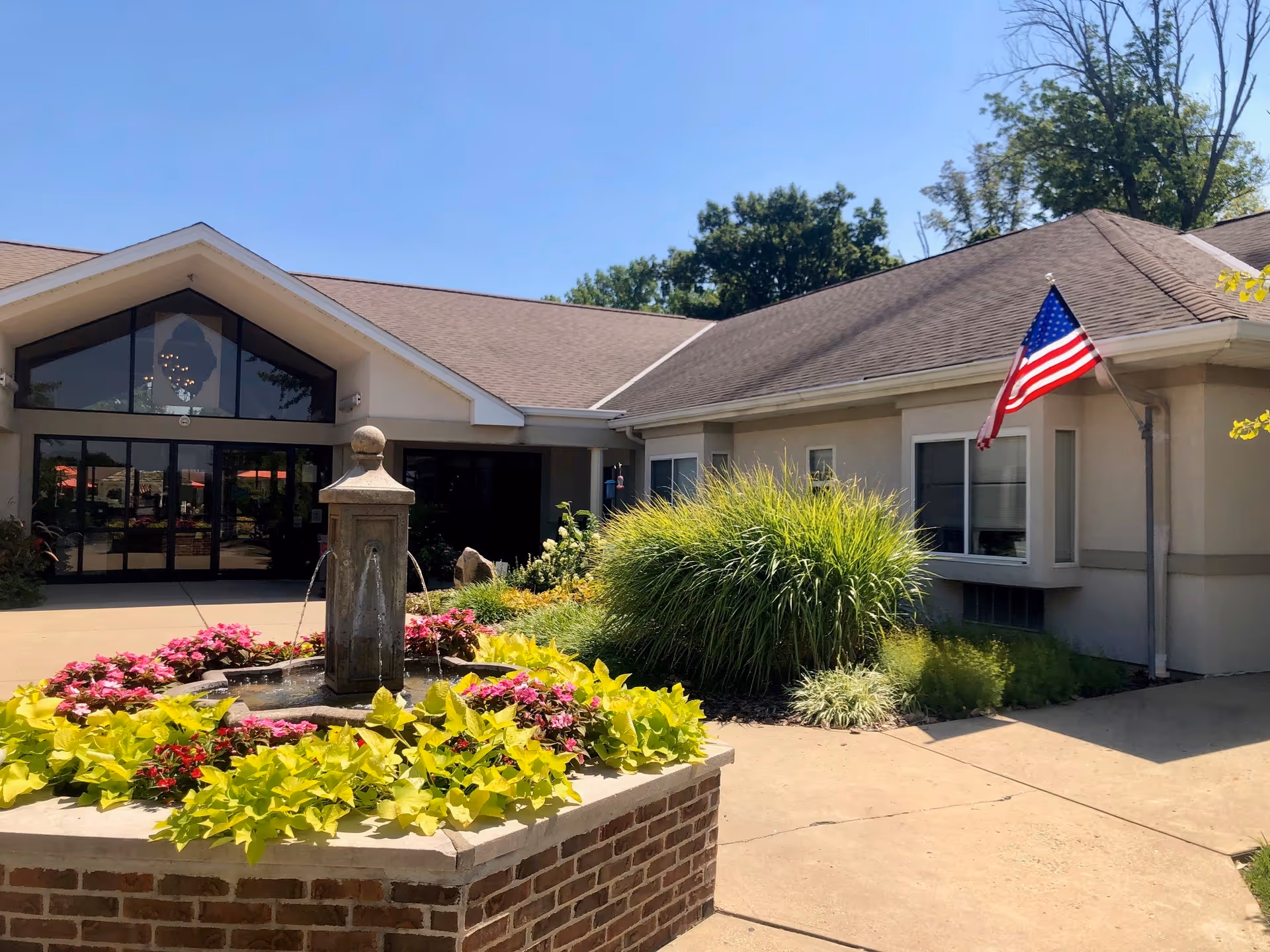 Front entrance of a one-story care facility with a central fountain surrounded by colorful flower beds, landscaping, and an American flag.