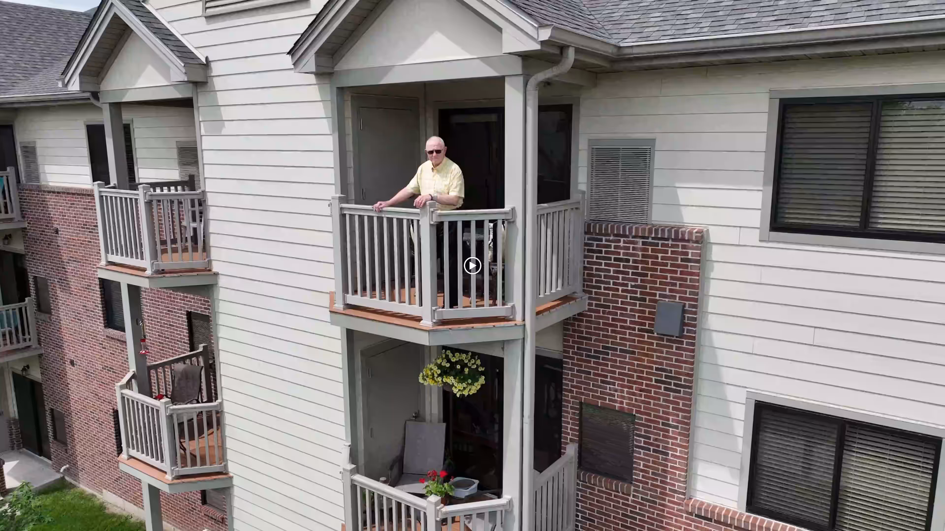 Exterior view of a multi-story residential building with balconies. An elderly man wearing sunglasses and a yellow shirt is standing on an upper balcony. The building features white siding with brick accents and several windows with closed blinds.