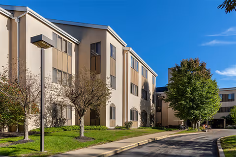 Exterior view of a multi-story senior living facility building with beige walls and multiple windows, surrounded by green grass, trees, and a paved driveway under a clear blue sky.