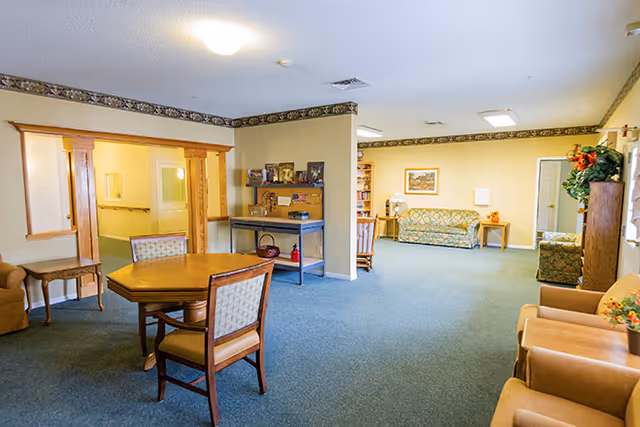 A spacious common area in an assisted living facility with a wooden table and chairs in the foreground, cushioned armchairs along the right side, and sofas against the far wall. The room has carpeted flooring, light yellow walls with decorative border trim, and various pieces of furniture including a small table with a lamp and a shelving unit with framed photos and decorations.