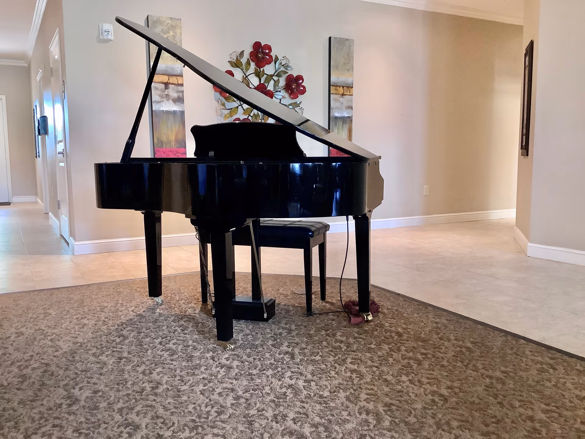 A black grand piano with a bench sits on a carpeted area in a neutral-toned interior common room with wall art behind it.