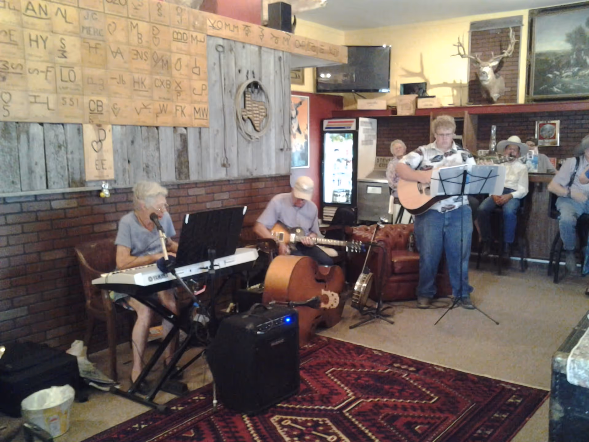 A group of elderly people playing musical instruments in a cozy room with rustic wooden and brick walls. One person is playing a keyboard, another is playing a guitar while seated, and a third person is standing and playing an acoustic guitar with a music stand in front. Several other elderly people are seated in the background, watching the performance. The room has a mounted deer head, a TV, and various decorations including a large wooden board with symbols and letters on the wall.