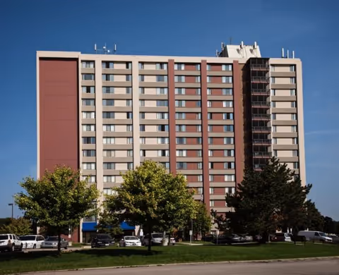 Exterior view of a multi-story residential building with beige and reddish-brown panels, several windows, and trees and parked cars in front under a clear blue sky.