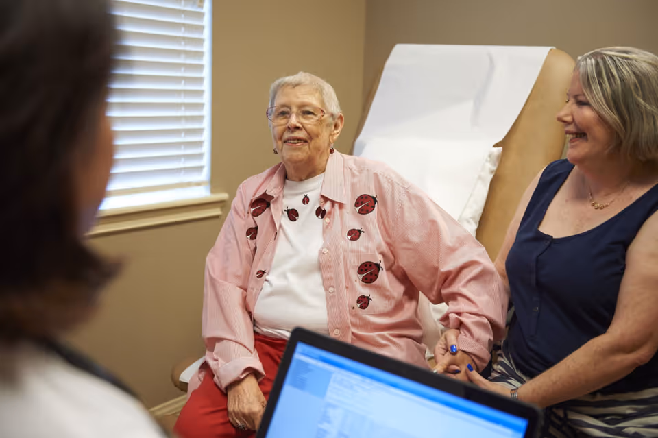 An elderly woman wearing a pink shirt with ladybug designs is sitting on a medical examination chair, holding hands with a smiling middle-aged woman beside her. A healthcare professional is partially visible in the foreground, holding a laptop with medical information on the screen. The setting appears to be a medical or consultation room with a window and beige walls.