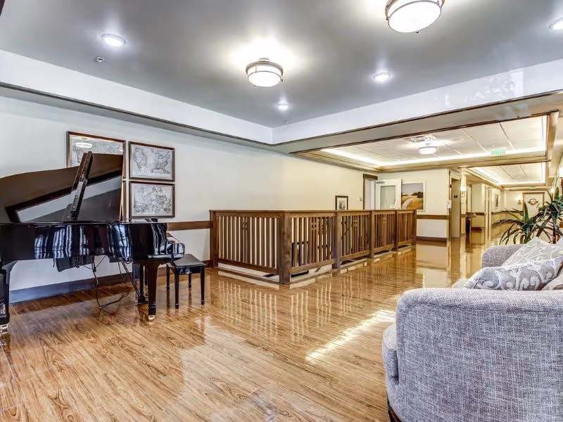 A spacious interior hallway with polished wooden floors, featuring a black grand piano with a matching bench on the left side. There are framed maps on the wall behind the piano. On the right side, there is a gray upholstered sofa with patterned cushions and a potted plant nearby. The ceiling has multiple round light fixtures, and wooden railings border a stairwell in the middle of the hallway.