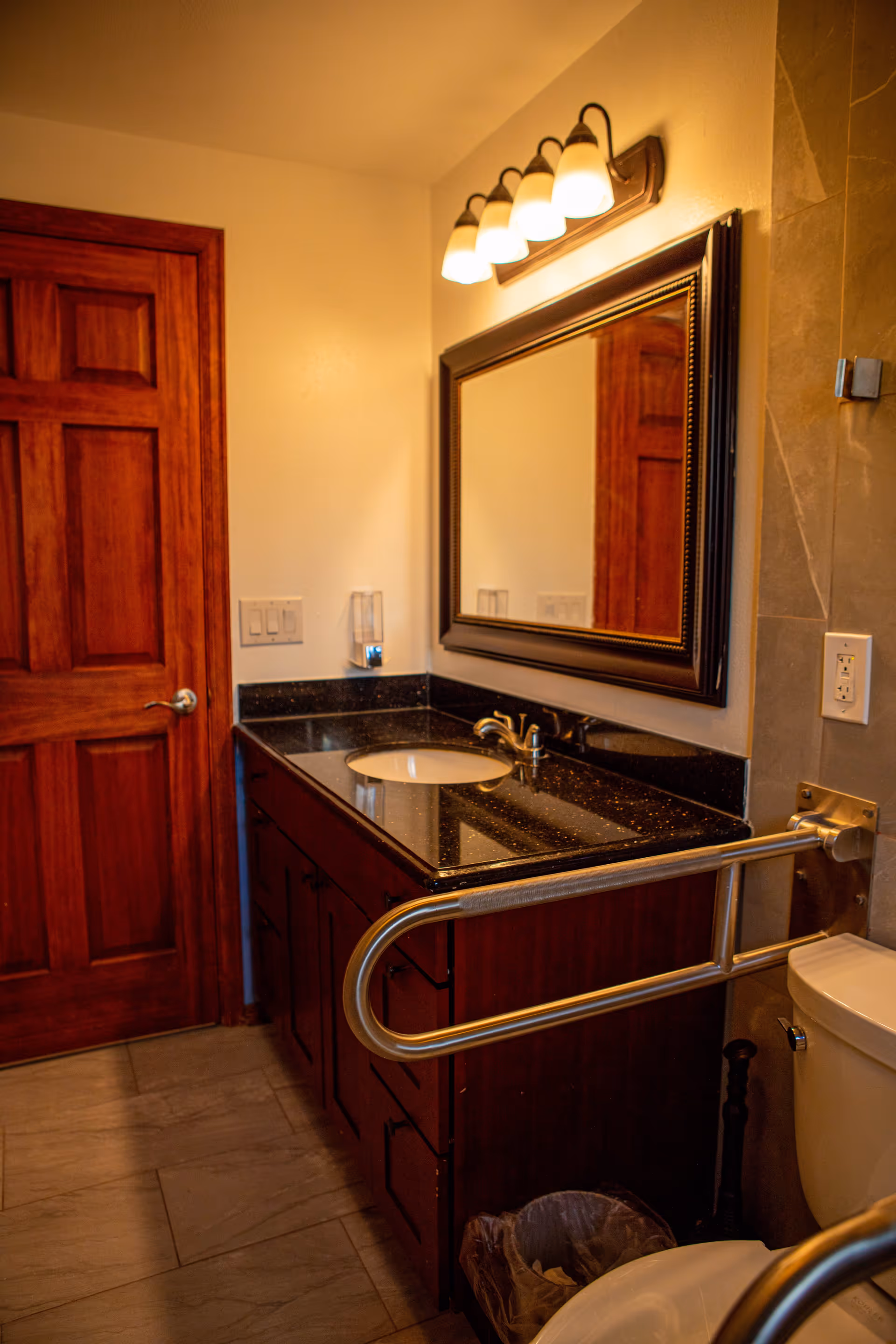 A bathroom interior featuring a wooden door, a dark granite countertop with a sink, a large framed mirror above the sink, a light fixture with five bulbs, a toilet with a metal grab bar, and a small trash bin on the floor.