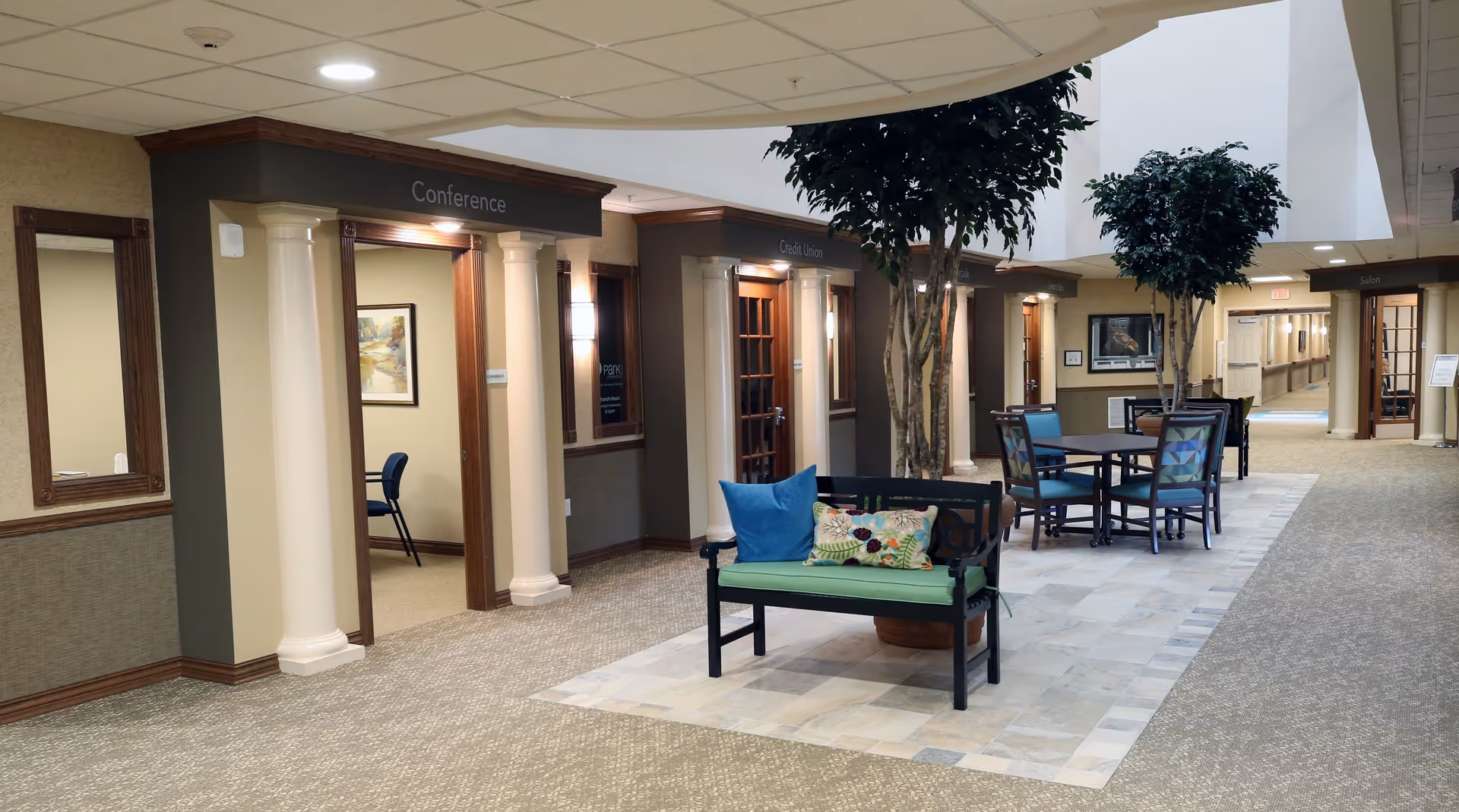 Interior hallway of Deerfield Springs Retirement Resort featuring a seating area with a bench and table with chairs, potted trees, and multiple doorways labeled Conference, Credit Union, and Salon. The space is well-lit with a combination of natural and artificial lighting.