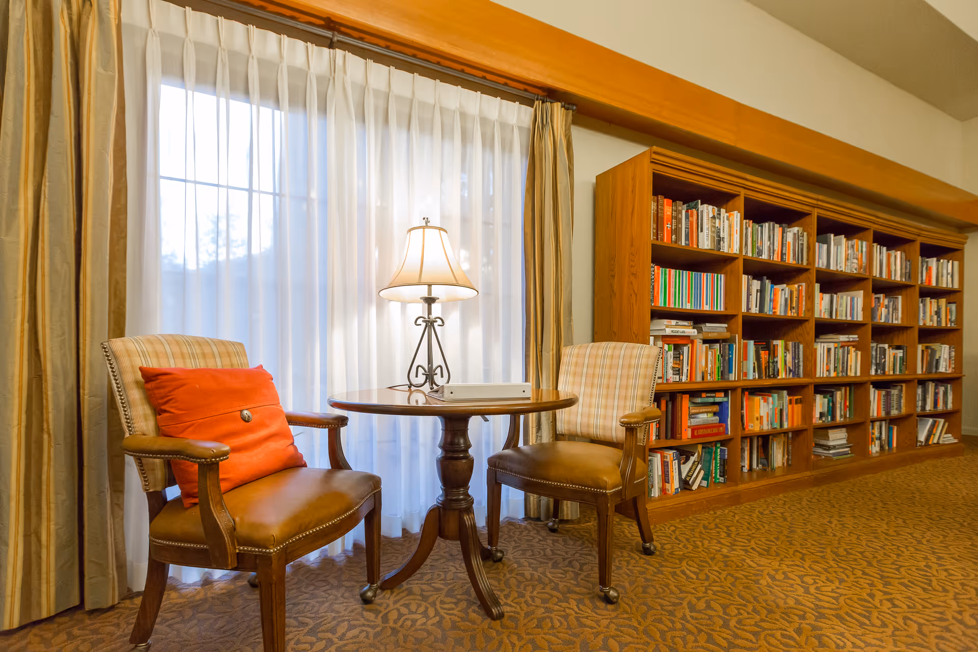 A cozy library seating area with two chairs, a round table, lamp, and tall bookshelf.