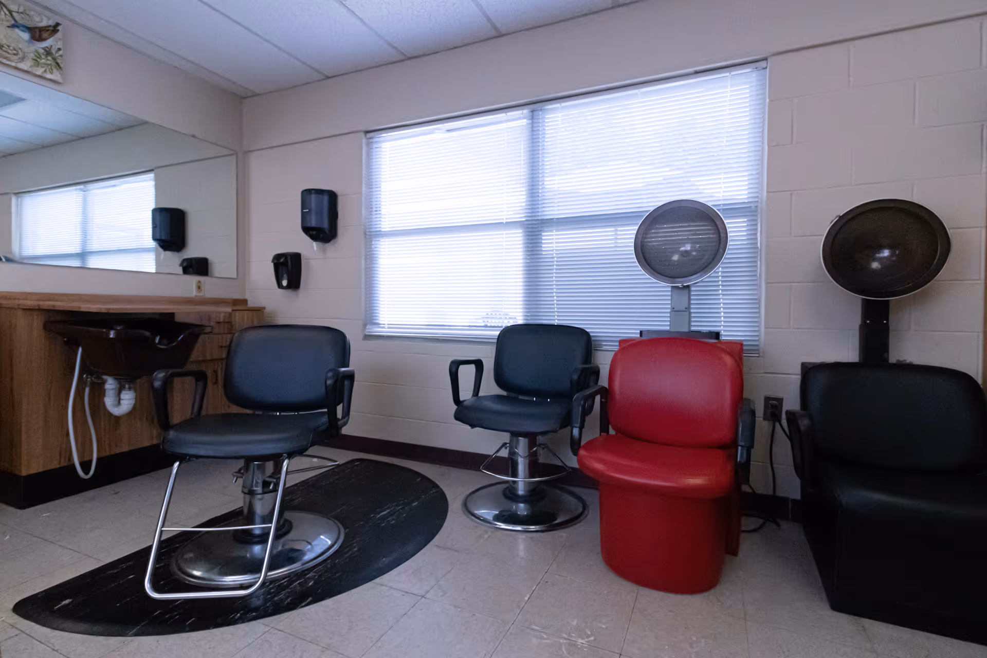 Interior view of a hair salon area with three salon chairs, two black and one red, positioned in front of hair dryers. There is a large mirror on the left wall above a wooden counter with a sink. The room has a window with blinds and light-colored walls.