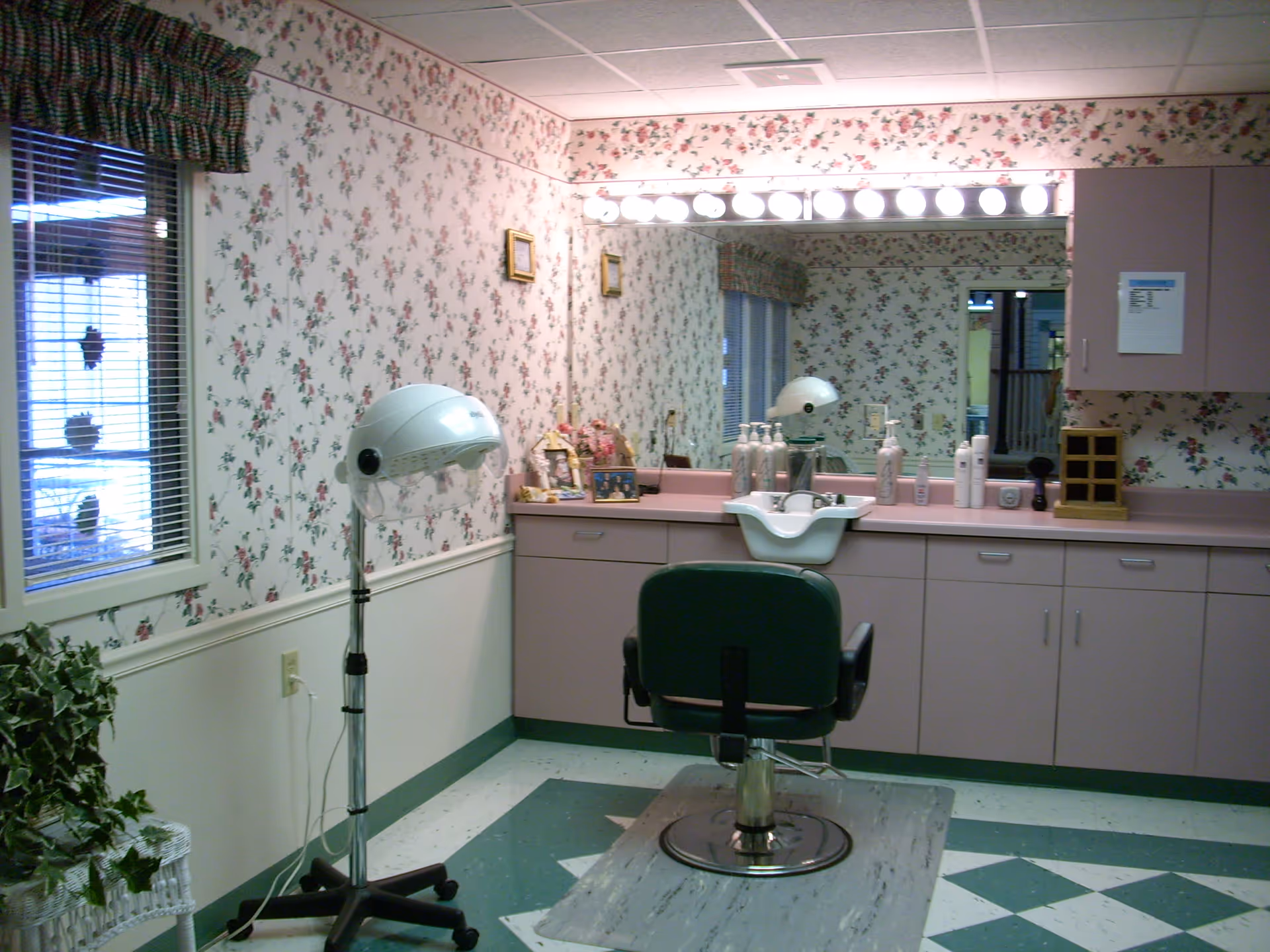 Small hair salon area with a styling chair, sink, hooded dryer, large mirror and floral wallpaper.