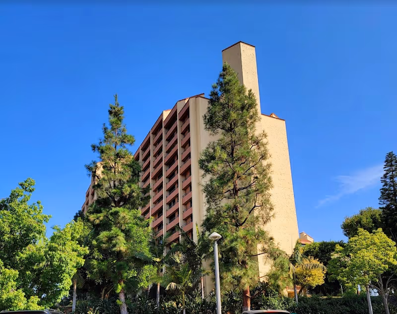 Tall multi-story residential tower surrounded by trees under a clear blue sky.