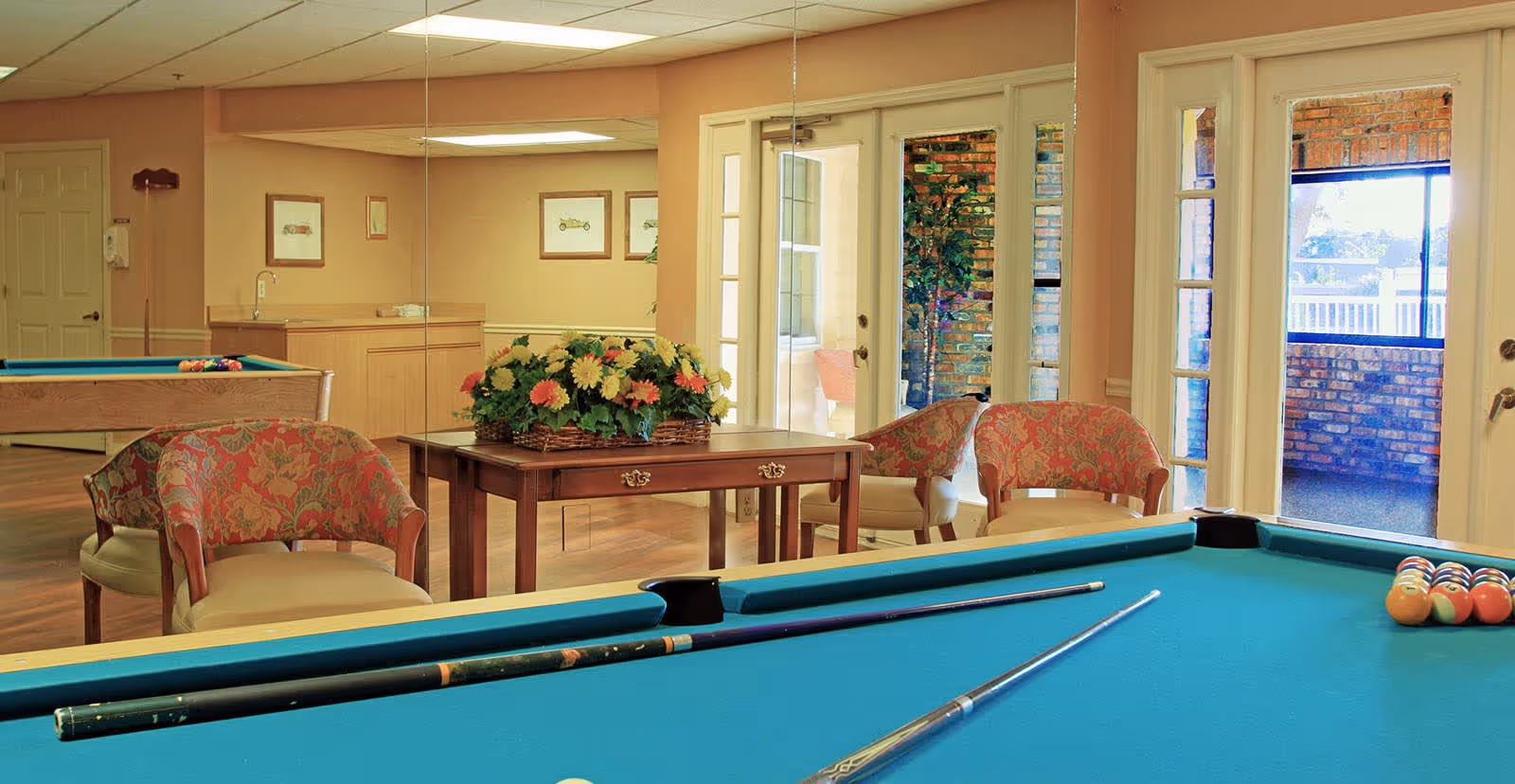 Interior lounge with a teal pool table in the foreground, floral armchairs and a table with a flower arrangement by mirrored walls and doors to an outdoor patio.