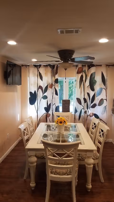 A dining room with a white rectangular table featuring a glass inset top, surrounded by six matching white chairs with patterned cushions. A small vase with yellow flowers is centered on the table. Behind the table, there are large windows covered with curtains that have a leaf pattern in shades of brown, green, and blue. A ceiling fan with a light fixture is mounted on the ceiling, and a flat-screen TV is mounted on the left wall. The floor is dark wood, and the room is softly lit by recessed ceiling lights.