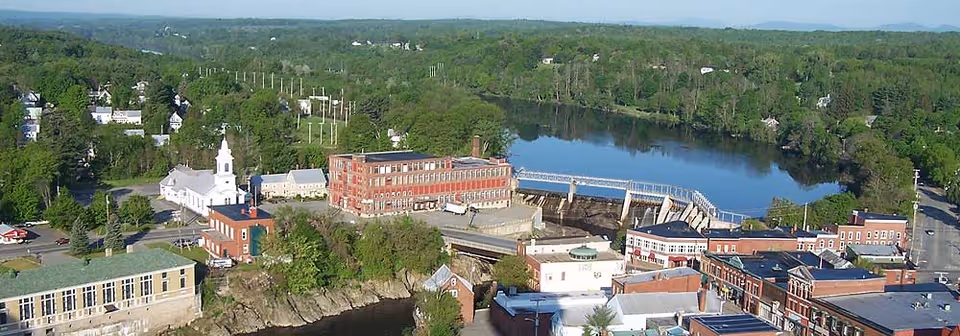 Aerial view of a small town with a river running through it, surrounded by green trees and hills in the background. The town features a mix of brick and white buildings, including a church with a steeple and a dam across the river.