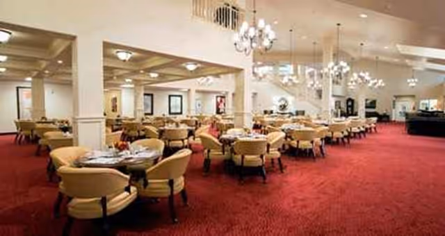 Spacious dining room with multiple round tables surrounded by beige upholstered chairs. The room features red carpet flooring, white walls, and several chandeliers hanging from the ceiling providing warm lighting. The area appears clean and well-organized, suitable for group dining.