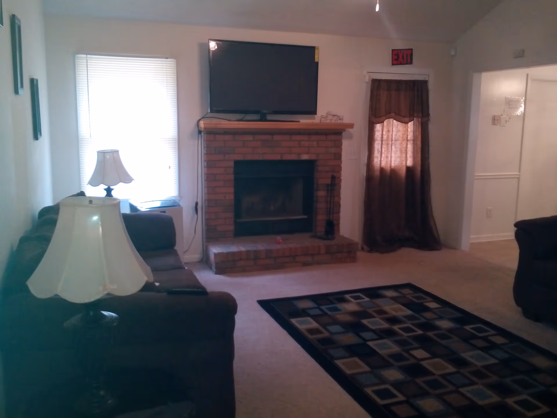 A living room with a brick fireplace and a flat-screen TV mounted above it. There is a window with blinds on the left side and a door with a curtain and an exit sign above it on the right. The room has two dark-colored sofas, a floor lamp with a white lampshade, and a patterned area rug on the carpeted floor.