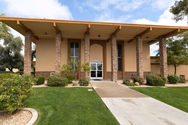 Front exterior view of Sunview Health And Rehabilitation Center building with a walkway leading to double glass doors, stone pillars, beige walls, and green landscaping with bushes and trees under a partly cloudy sky.