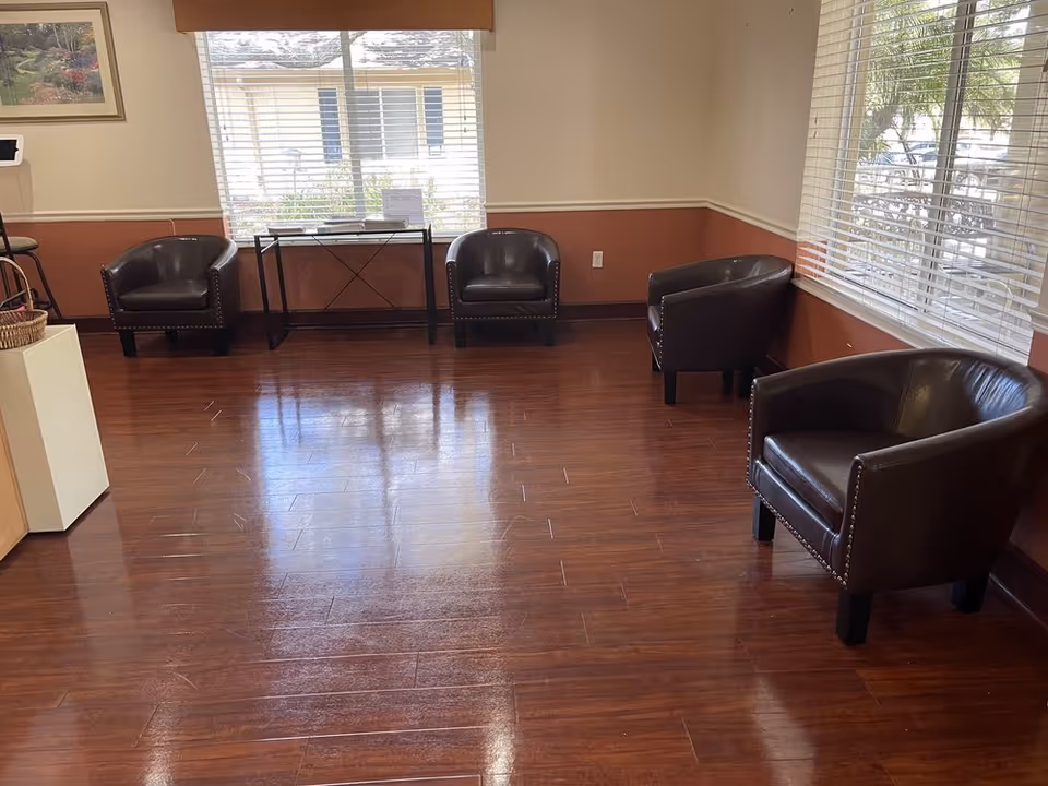 Sunlit waiting area with five brown leather armchairs along the walls, wood flooring, and large windows.