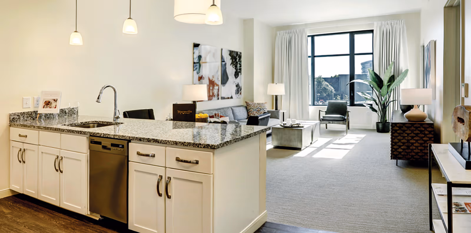 Open-plan apartment interior featuring a granite kitchen island in the foreground and a bright living room with seating and a large window in the background.