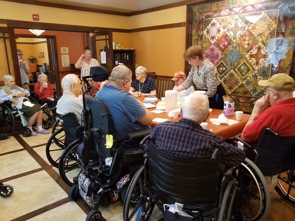 Elderly residents in wheelchairs gathered around a table in a communal dining/activity room while a staff member assists them.
