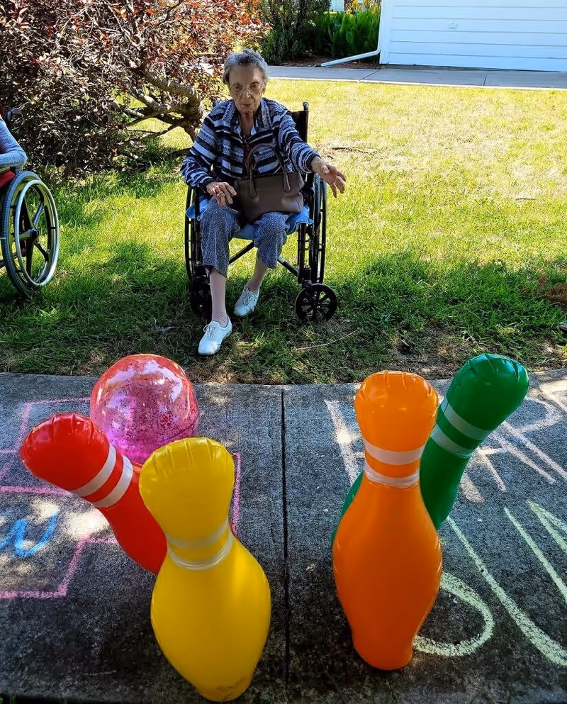 An elderly woman in a wheelchair outdoors on a sunny day, playing a game of inflatable bowling with colorful pins and a pink ball on a concrete surface with chalk drawings.