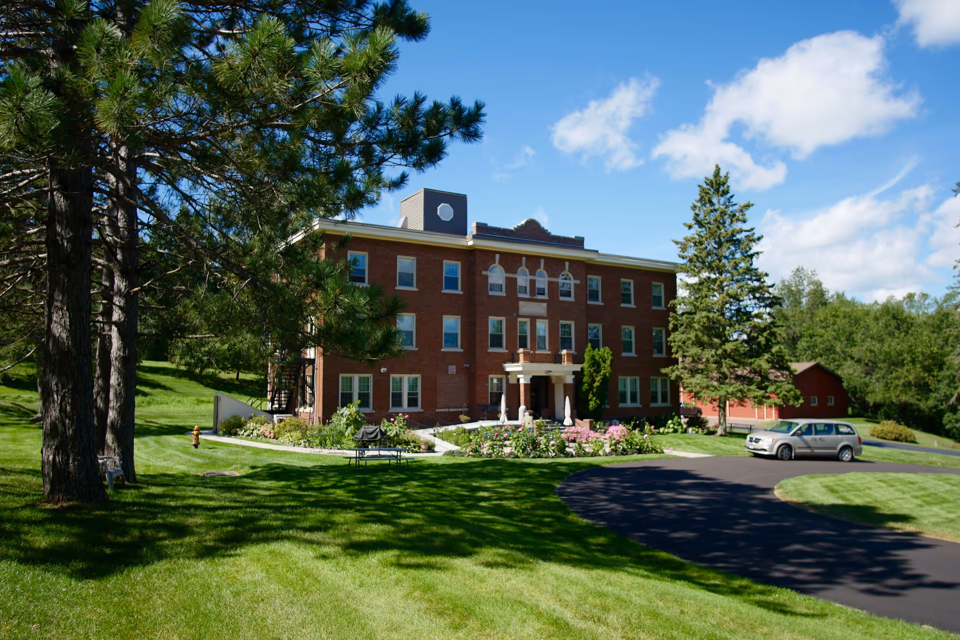 A three-story brick building with white trim surrounded by green grass, trees, and a clear blue sky. There is a paved driveway with a parked silver car and a garden with flowers in front of the building.