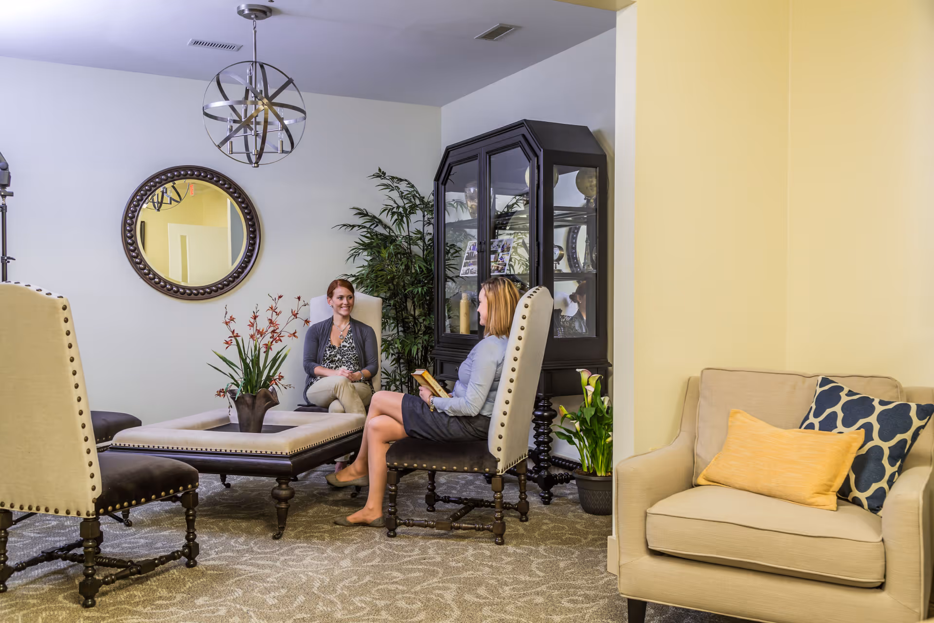 Two women sitting and conversing in a well-decorated living room area with beige and dark wood furniture, a large mirror on the wall, a black display cabinet, plants, and a beige armchair with decorative pillows.