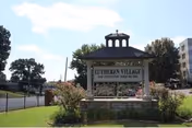 Outdoor view of a sign for Lutheran Village, a senior living community, situated on a grassy area with bushes and trees nearby. The sign is mounted on a stone base with a small roof structure above it. A building and a fence are visible in the background under a partly cloudy sky.