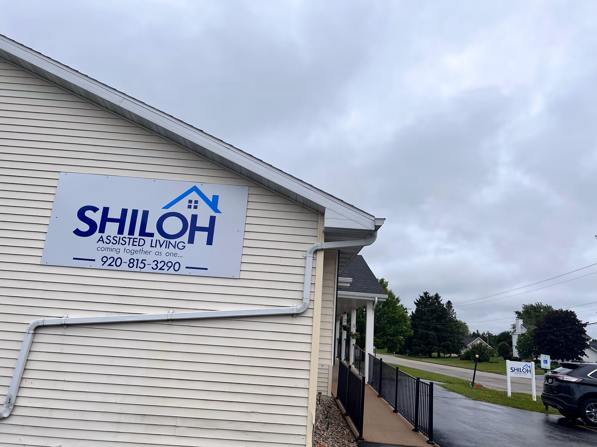 Exterior view of the Shiloh Assisted Living building with a large sign on beige siding, a walkway and parking area under a cloudy sky.