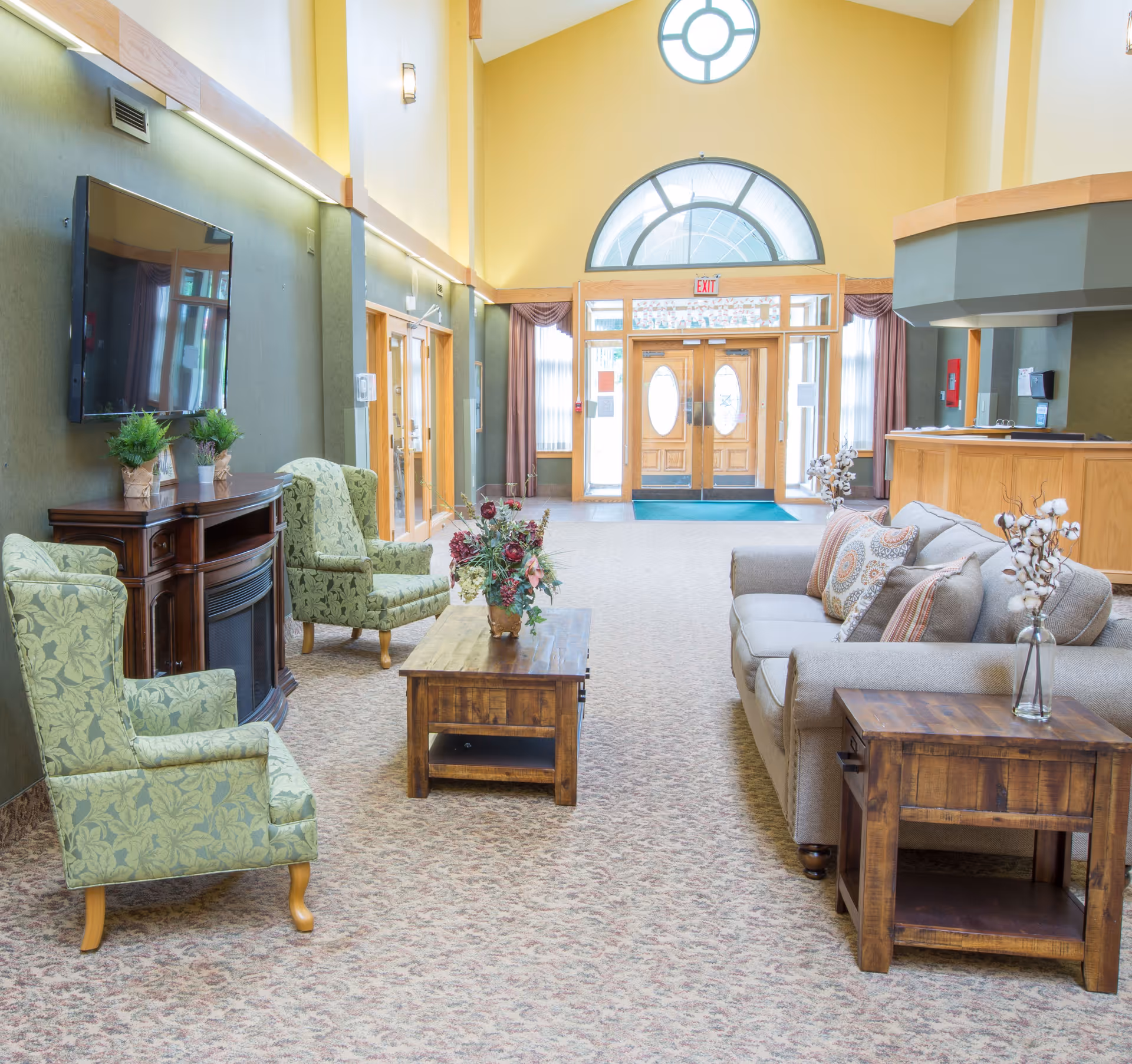 Sunlit senior living lobby with sofas and armchairs around a wooden coffee table, a wall-mounted TV, and a reception desk near double front doors.