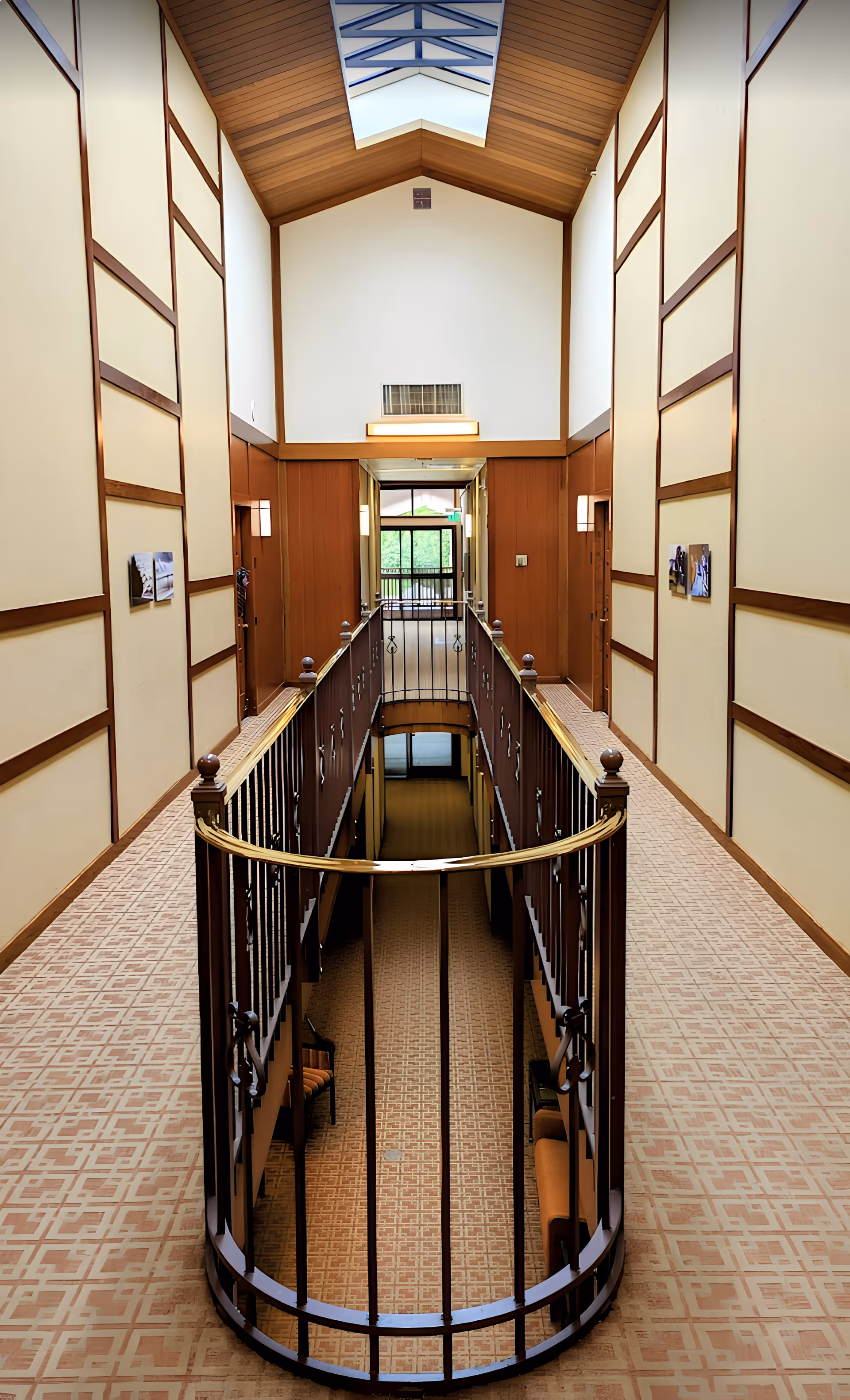 Interior hallway of Spring Lake Village with a patterned carpet, wooden panel walls, and a skylight ceiling. The hallway has a railing around an open central area that looks down to a lower floor with seating visible below. Doors and wall-mounted lights line the corridor.