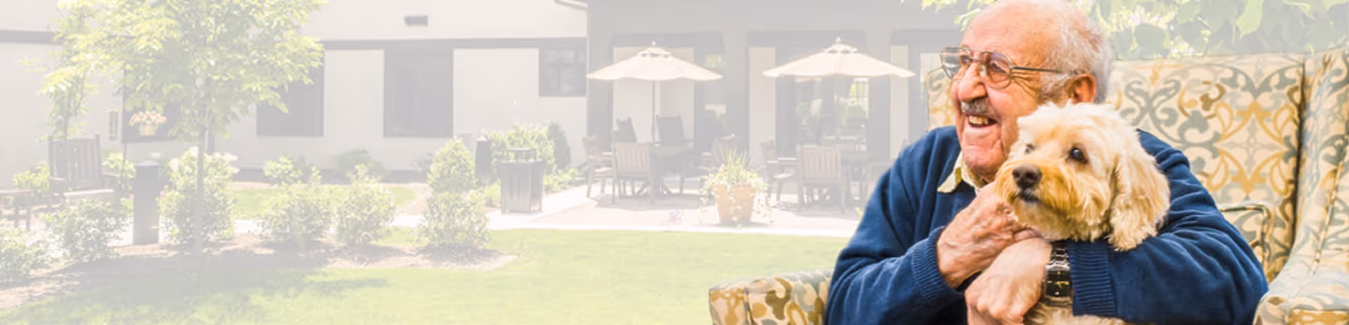 An elderly man with glasses sitting on a patterned armchair outdoors, smiling and hugging a small light brown dog. In the background, there is a garden area with green grass, bushes, and patio tables with umbrellas in front of a building.