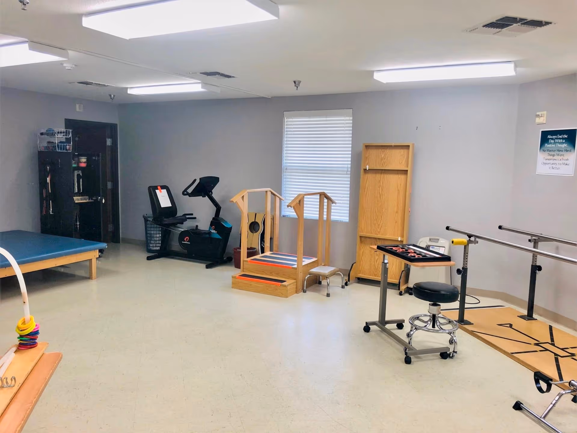 A physical therapy room with a recumbent exercise bike, parallel bars, wooden therapy steps, treatment table and stools.