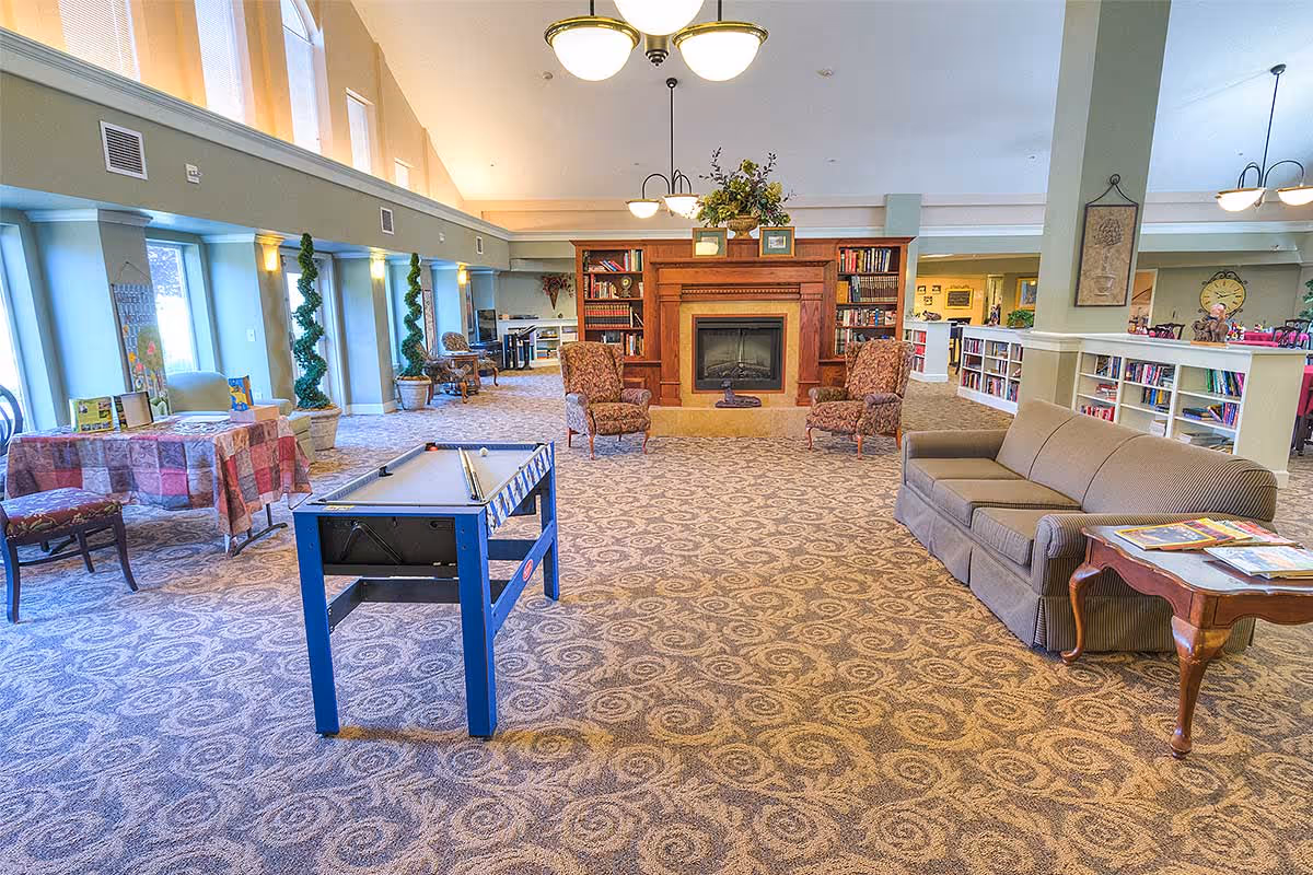A spacious senior living common area with patterned carpet, a blue foosball table, a brown sofa with a side table holding magazines, two upholstered armchairs facing a wooden fireplace with built-in bookshelves, and large windows letting in natural light.