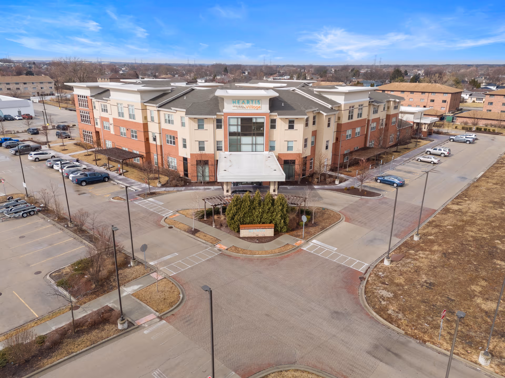 Aerial view of a large, multi-story senior living facility building with a covered entrance and surrounding parking lots. The building has a beige and brick exterior with multiple windows. There are landscaped areas with trees and shrubs near the entrance and parking areas. The sky is clear with some clouds.