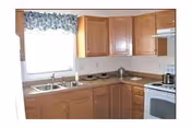 A kitchen with wooden cabinets, a double sink under a window with a floral valance, a white stove with an oven, and a countertop with a few items on it.