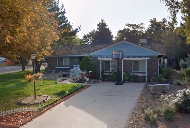 Single-story residential building with a brick and blue exterior, a driveway with a portable basketball hoop, surrounded by trees and landscaping.